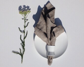 May include: A white ceramic napkin ring holding a folded brown linen napkin on a white plate. A sprig of white flowers is on the left side of the image.