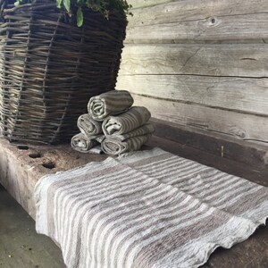 May include: A stack of four brown and white striped linen towels on a wooden surface. The towels are folded and stacked neatly. A wicker basket is in the background.