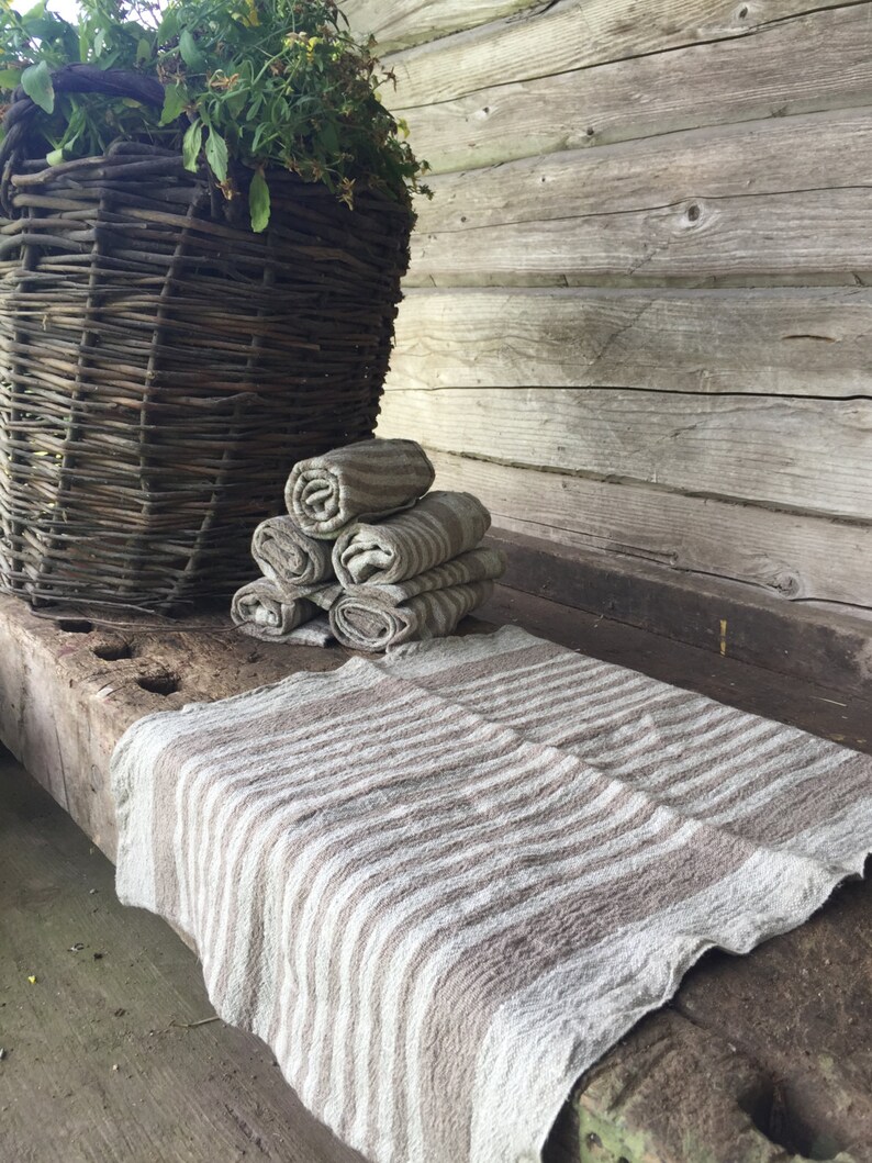 May include: A stack of four brown and white striped linen towels on a wooden surface. The towels are folded and stacked neatly. A wicker basket is in the background.