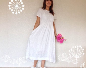 Puede incluir: Un vestido blanco maxi con mangas cortas y cintura fruncida. El vestido está hecho de una tela ligera y tiene una silueta fluida. La modelo lleva zapatillas blancas y sostiene un ramo de flores rosas.