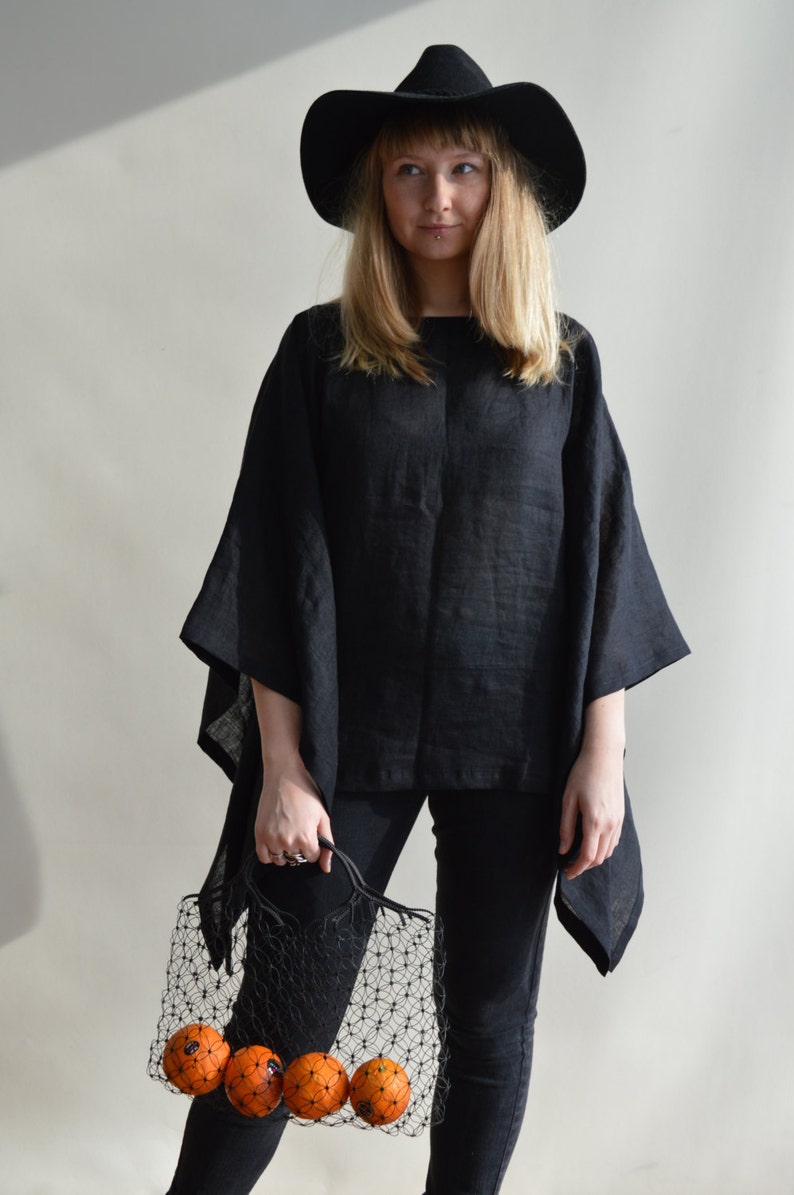 May include: A woman wearing a black wide-brimmed hat and a black linen top holds a black mesh bag filled with oranges.