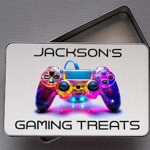 May include: A rectangular metal tin with a lid, featuring a colourful, illuminated video game controller design. The text "JACKSON'S GAMING TREATS" is printed in black on the lid. The controller has a neon colour scheme with blue, purple, yellow, and pink hues.