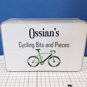 May include: A white rectangular storage tin with the text "Ossian's Cycling Bits and Pieces" and a bicycle graphic in green and black. The tin has a silver trim. The background is blue.