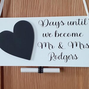 May include: White rectangular sign with a black heart and the text "Days until we become Mr & Mrs Rodgers". A small scroll is attached below the sign with a black and white ribbon. White ribbon is used for hanging.