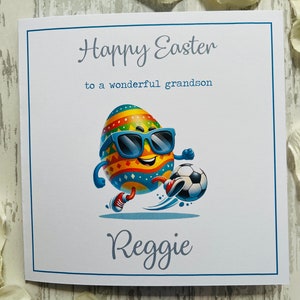 May include: A white Easter card with a blue border. The card features a cartoon egg wearing sunglasses and playing football. Text reads "Happy Easter to a wonderful grandson" and the name "Reggie".