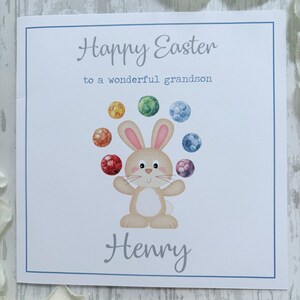 May include: A white Easter card with a blue border. The card features the text "Happy Easter to a wonderful grandson" and a cartoon bunny juggling colorful balls. The name "Henry" is written below the bunny.