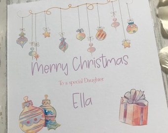 May include: A square Christmas card with the words "Merry Christmas" in purple script. The card features watercolor illustrations of hanging ornaments, a gift, and the text "To a special Daughter Ella."