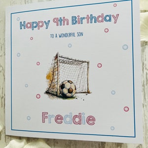 May include: A white birthday card with a blue border. The card features the text "Happy 9th Birthday to a Wonderful Son" in red and blue. A football is in front of a net. The name "Freddie" is written below the image.