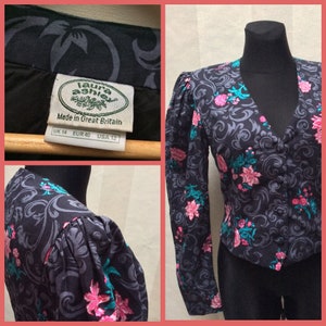 May include: A black and grey floral print blouse with puffed sleeves. The blouse has a V-neckline and a button closure. The label reads "Laura Ashley" and "Made in Great Britain". The size tag reads "UK 14, EUR 40, USA 12".