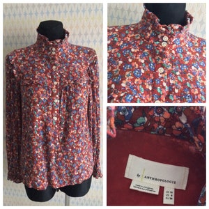 Pu&ograve; includere: Una blusa rossa a maniche lunghe con stampa floreale e colletto alto. La blusa ha una chiusura con bottoni sul davanti e un colletto arricciato. L'etichetta riporta "by Anthropologie" e include la taglia: UK 10, US 6, EU 38.
