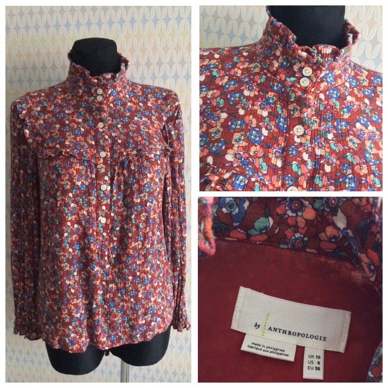Pu&ograve; includere: Una blusa rossa a maniche lunghe con stampa floreale e colletto alto. La blusa ha una chiusura con bottoni sul davanti e un colletto arricciato. L'etichetta riporta "by Anthropologie" e include la taglia: UK 10, US 6, EU 38.