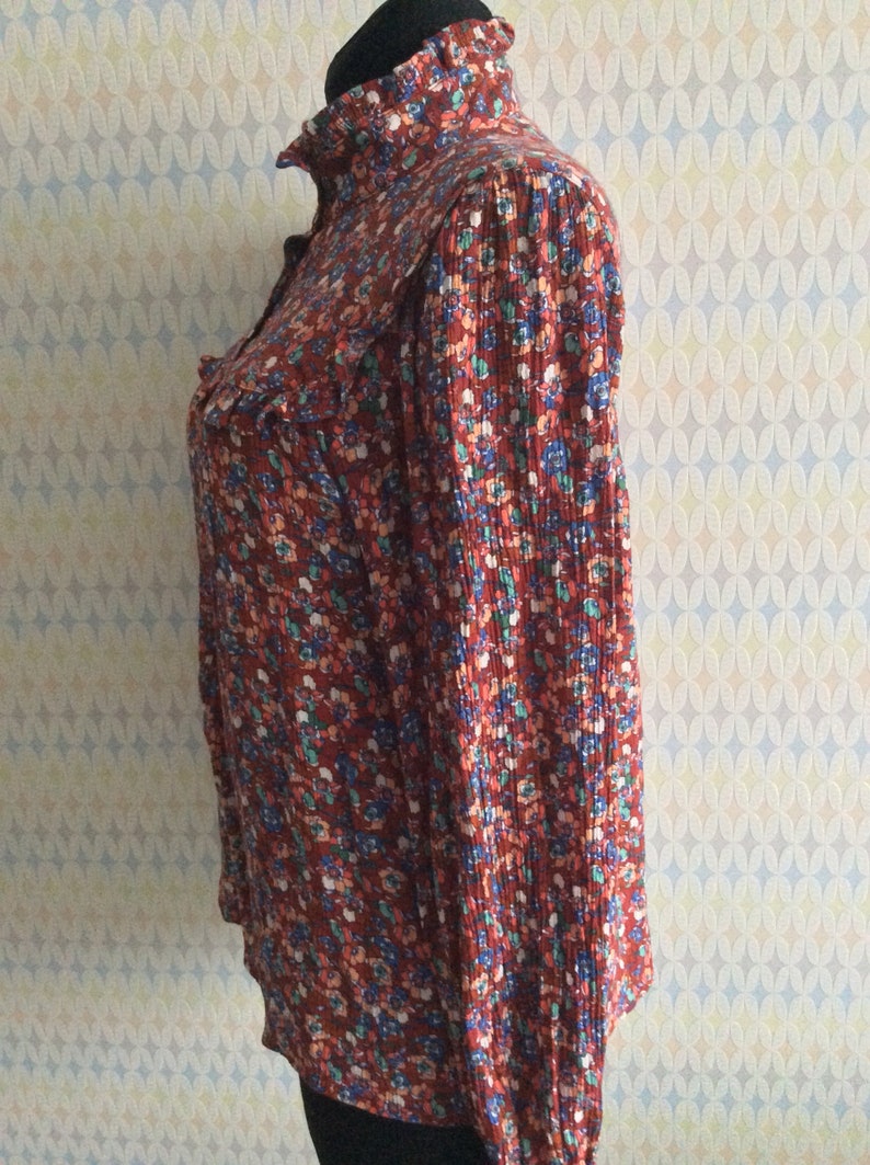Pu&ograve; includere: Una blusa a maniche lunghe color ruggine con colletto a volant e stampa floreale in tonalit&agrave; blu, rosa e verde.
