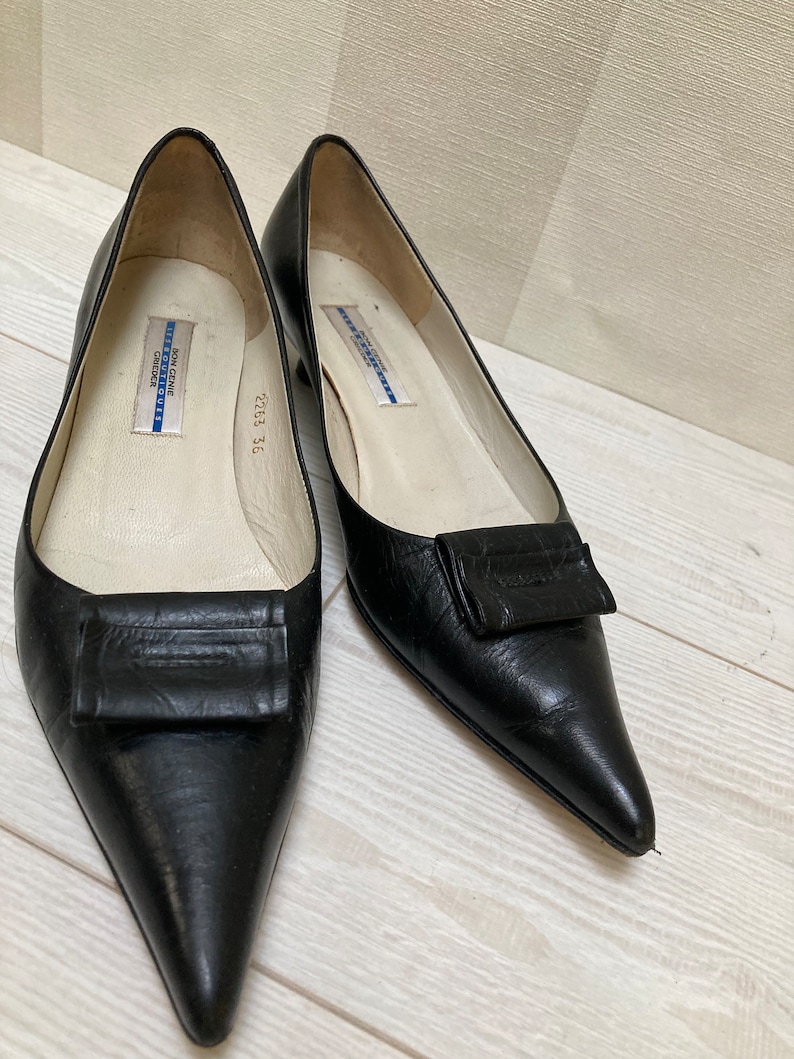 May include: A pair of black leather pointed-toe shoes with a small heel and a decorative buckle on the vamp. The shoes are labeled "Don Genie Boutique" and "2253 36".