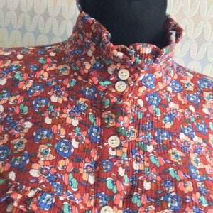 Pu&ograve; includere: Una blusa con stampa floreale rosso, bianco e blu con un colletto alto arricciato e chiusura con bottoni.