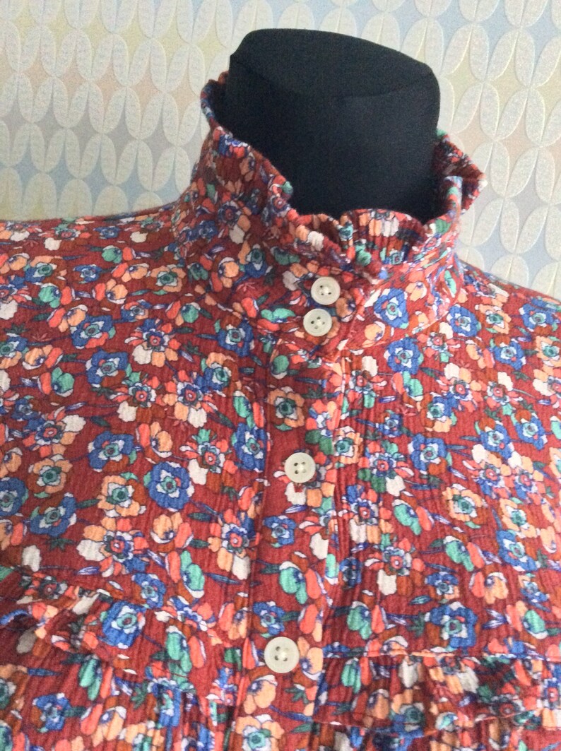 Pu&ograve; includere: Una blusa con stampa floreale rosso, bianco e blu con un colletto alto arricciato e chiusura con bottoni.