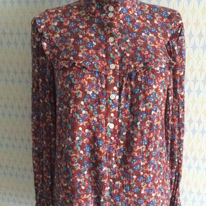 Pu&ograve; includere: Una blusa a maniche lunghe con stampa floreale rosso, bianco e blu, con colletto alto e chiusura con bottoni.