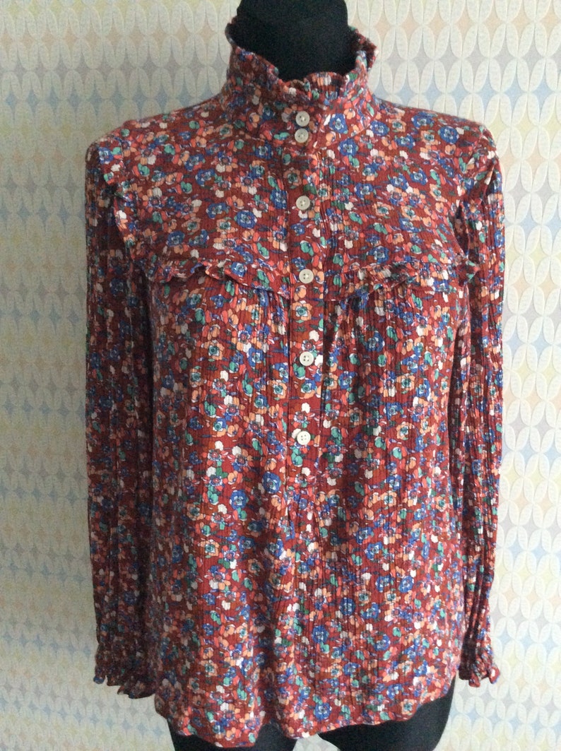 Pu&ograve; includere: Una blusa a maniche lunghe con stampa floreale rosso, bianco e blu, con colletto alto e chiusura con bottoni.