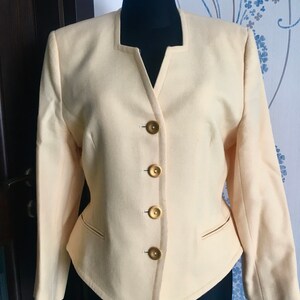 May include: A cream-colored blazer with a V-neck and five gold buttons. The blazer has a fitted silhouette and long sleeves.