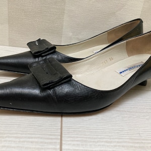 May include: Black leather heeled shoes with a large black bow on the front. The shoes have a white insole with blue text that reads "Made in Italy 36".