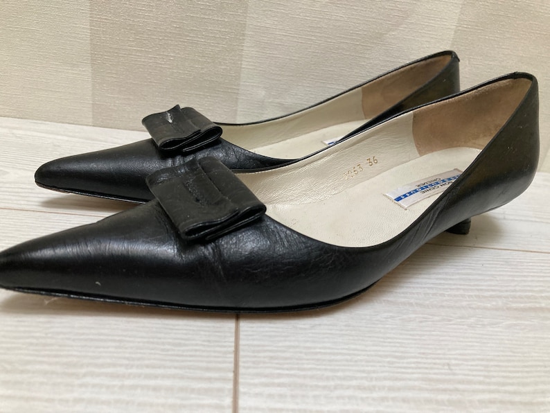 May include: Black leather heeled shoes with a large black bow on the front. The shoes have a white insole with blue text that reads "Made in Italy 36".