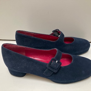 May include: A pair of navy blue suede shoes with a buckle fastening and a small heel. The shoes have a red lining.