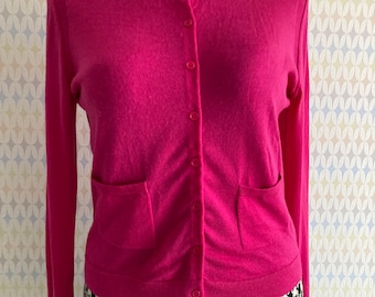 May include: A pink long-sleeved cardigan with a button-up front and two pockets. The cardigan is on a mannequin and is being displayed against a patterned background.