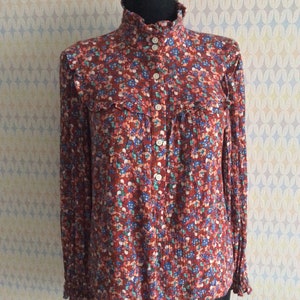 Pu&ograve; includere: Una blusa a maniche lunghe con stampa floreale rosso, bianco e blu, con colletto alto e chiusura con bottoni.