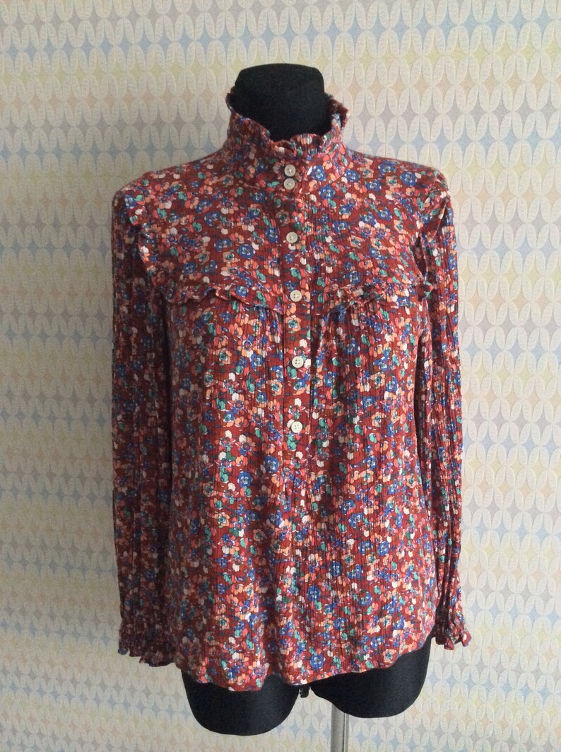 Pu&ograve; includere: Una blusa a maniche lunghe con stampa floreale rosso, bianco e blu, con colletto alto e chiusura con bottoni.