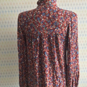 Pu&ograve; includere: Una blusa a maniche lunghe con stampa floreale rossa, blu e verde, collo alto e polsini con volant.