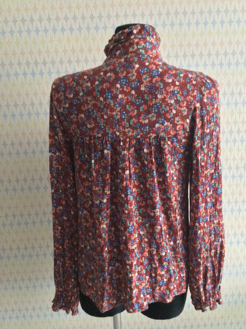 Pu&ograve; includere: Una blusa a maniche lunghe con stampa floreale rossa, blu e verde, collo alto e polsini con volant.