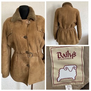 Puede incluir: Una chaqueta de ante marrón con cuello de piel de oveja y cintura ajustada. La chaqueta tiene cierre de botones y dos bolsillos de solapa. La etiqueta dice "Baily's of Glastonbury" y presenta un logotipo de oveja. La etiqueta de talla indica una talla 16 y una longitud de 97 cm.