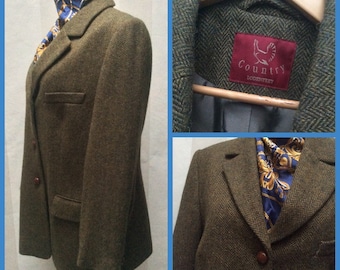 May include: A green tweed blazer with a herringbone pattern. The blazer has a single breasted closure with two buttons. The label on the inside of the blazer reads "Country Lodensfrey".