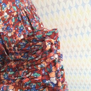 Pu&ograve; includere: Primo piano di un tessuto a stampa floreale rosso, blu e verde con un dettaglio arricciato. Il tessuto &egrave; morbido e ha un aspetto vintage.