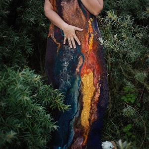 Può includere: Una donna indossa un lungo abito fluido in lana feltrata. L'abito è decorato con motivi astratti in tonalità di blu, arancio e giallo. La donna è in piedi in un ambiente boschivo.