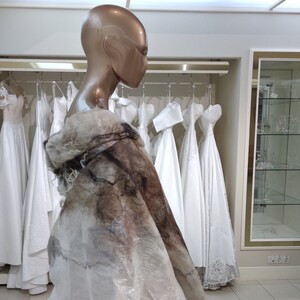 May include: A mannequin wearing a white bridal shawl with a brown and gray pattern. The shawl is draped over the mannequin's shoulders and falls to the floor. The mannequin is standing in a room with several white wedding dresses hanging on racks.