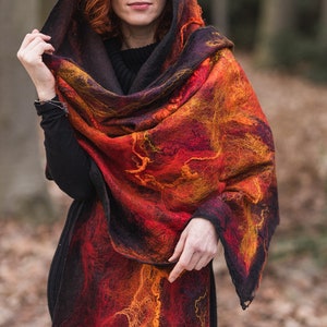 May include: A hooded shawl in fiery shades of red, orange, and black. The felted wool garment features a textured, abstract design. The shawl is draped over the shoulders and head, creating a warm and stylish accessory.