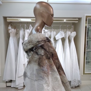 May include: A mannequin wearing a white and brown shawl-like garment, standing in front of a rack of white wedding dresses.
