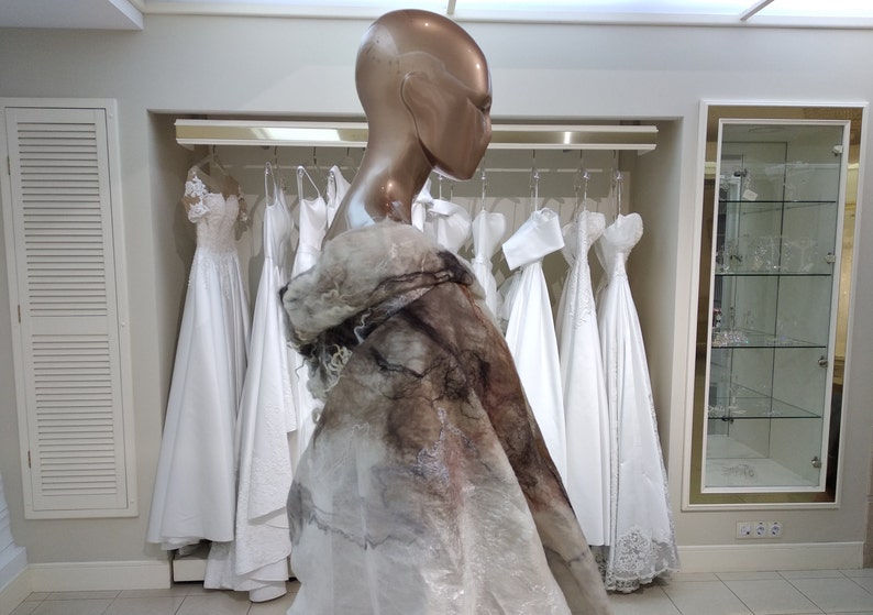 May include: A mannequin wearing a white and brown shawl-like garment, standing in front of a rack of white wedding dresses.