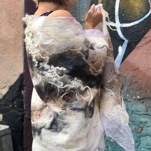 May include: A white, brown, and black felted wool scarf with a flowing, abstract design. The scarf is draped over a person's shoulders.