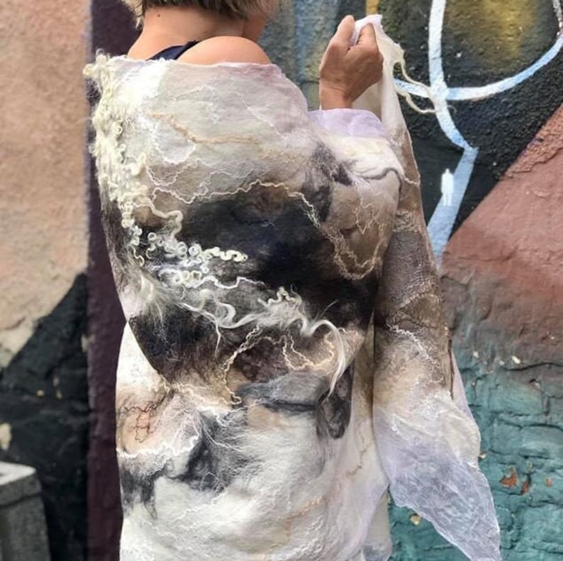 May include: A white, brown, and black felted wool scarf with a flowing, abstract design. The scarf is draped over a person's shoulders.