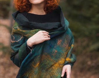May include: A felted wool shawl in shades of teal, green, and gold. The shawl has a hooded design and is draped over the shoulders. The texture of the wool is visible, with a mix of colors creating a unique pattern.