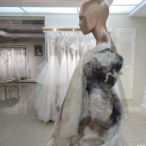 May include: A white wedding dress with a long, flowing skirt and a fitted bodice. The dress is made of a soft, flowing fabric and has a delicate lace trim. The dress is displayed on a mannequin in a bridal shop.