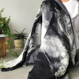Puede incluir: Una bufanda de lana afieltrada en tonos grises y negros, con detalles blancos. La bufanda presenta un diseño veteado con un patrón fluido en forma de nube. Los bordes tienen un aspecto texturizado y con flecos. La bufanda está drapeada sobre los hombros de una persona.