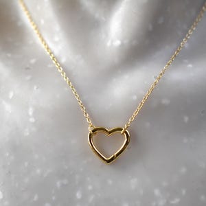 May include: A gold-colored necklace featuring a delicate open heart pendant. The pendant is suspended from a fine chain. The necklace is displayed against a soft, blurred background, highlighting the jewelry's design.