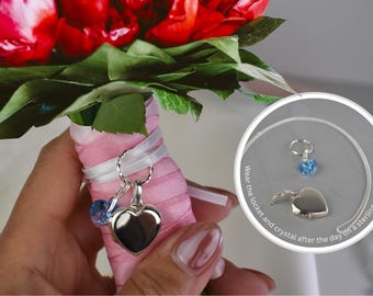 May include: A bouquet of red flowers with green leaves, wrapped in pink ribbon and white satin. A silver heart-shaped locket and blue crystal charm hang from the bouquet. A small, round image shows the locket and crystal on a silver chain.