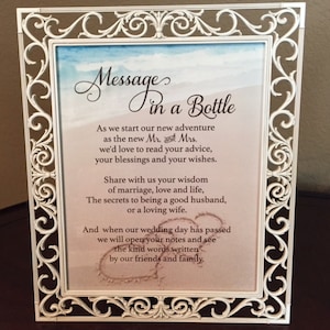 May include: A white, ornate picture frame with the words "Message in a Bottle" and wedding-related text. The frame has a decorative, swirling design. The background shows a beach scene with a heart drawn in the sand.