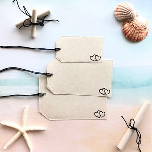 May include: Three cream-colored gift tags with black heart outlines and black string. Two rolled scrolls tied with black string, a white starfish, and two seashells are also present. The background has a beach theme.