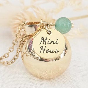 Może przedstawiać: Złoty naszyjnik z okrągłą zawieszką i małą zieloną koralikiem. Na zawieszce widnieje napis "Mini Nous". Naszyjnik leży na białej powierzchni.
