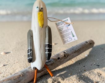 Puede incluir: Una figura de gaviota hecha de madera flotante de color claro con un pico amarillo pintado, ojos azules y alas grises con rayas blancas y negras. La figura tiene patas naranjas y una etiqueta de papel blanco atada con una cuerda que dice Kemzma Designz.