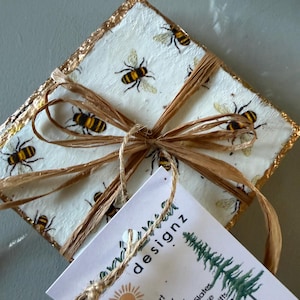 May include: Square coaster set with a bee pattern, tied with raffia. The coasters have a white background with gold trim and are decorated with black and yellow bees. A tag with the word "designz" is attached.